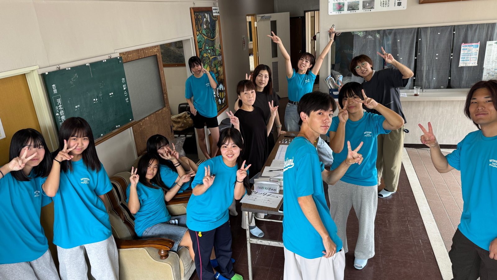 外日角公民館の受付で、メンバーとスタッフがピースサインをする様子